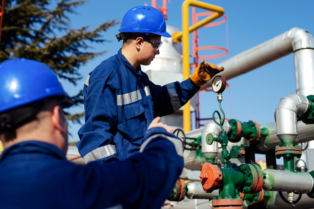 Two engineers in blue coveralls checking a pressure gauge on refinery pipework